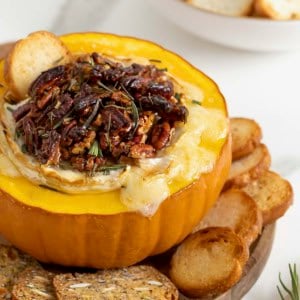 Baked brie in a hollowed out mini pumpkin topped with chopped pecans and rosemary on a wooden board on white marble counter. There are crackers and crostini on the board surrounding the pumpkin.