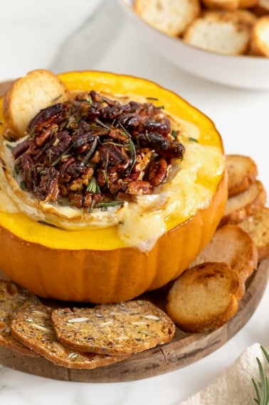 Baked brie in a hollowed out mini pumpkin topped with chopped pecans and rosemary on a wooden board on white marble counter. There are crackers and crostini on the board surrounding the pumpkin.