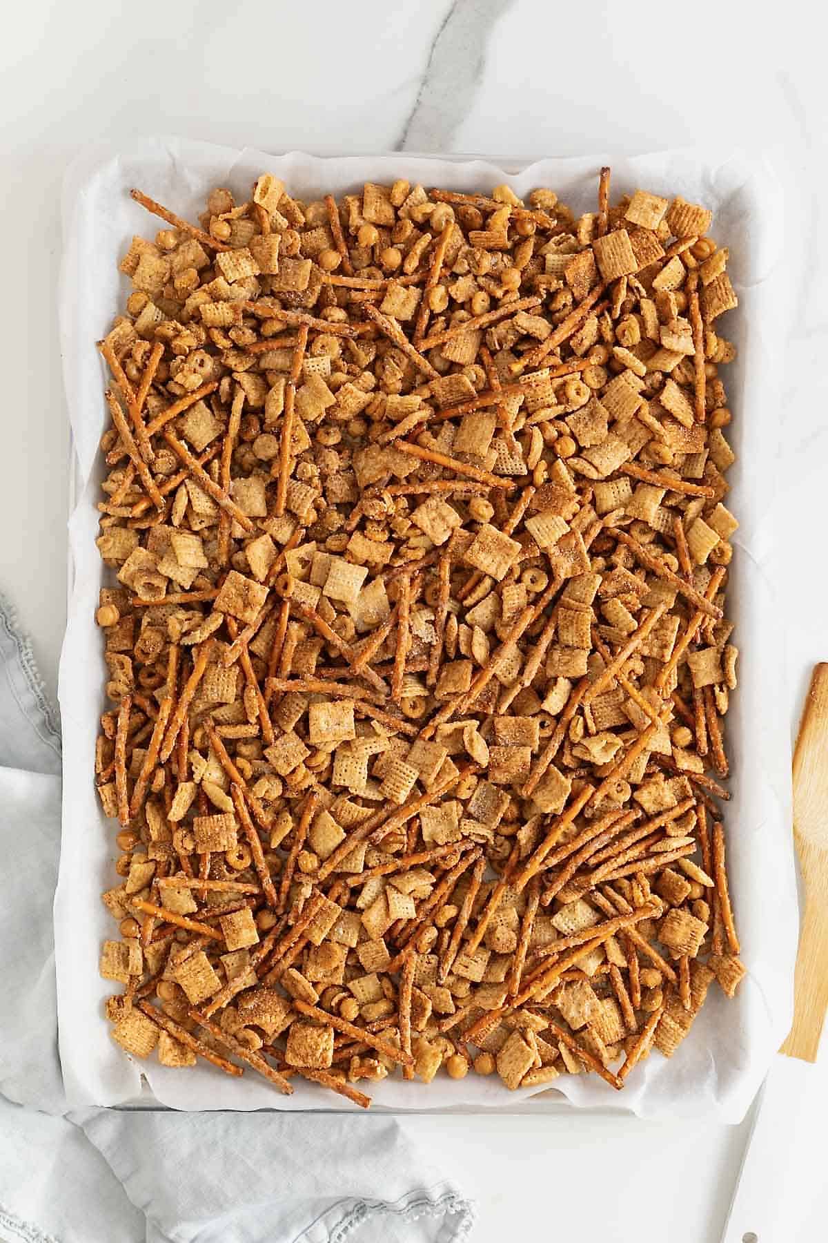 A parchment lined sheet pan of churro snack mix on a white surface. There is a wooden spoon to the right of the sheet pan.