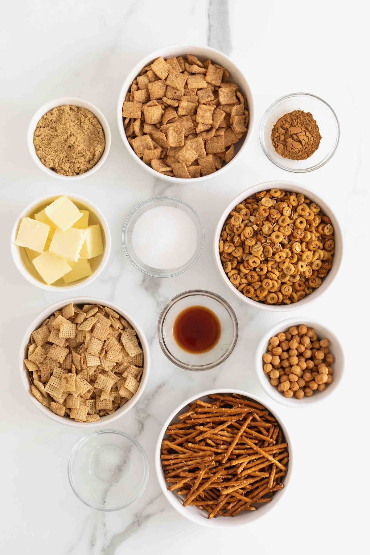 Ingredients to make churro snack mix in small glass dishes on a white marble counter.