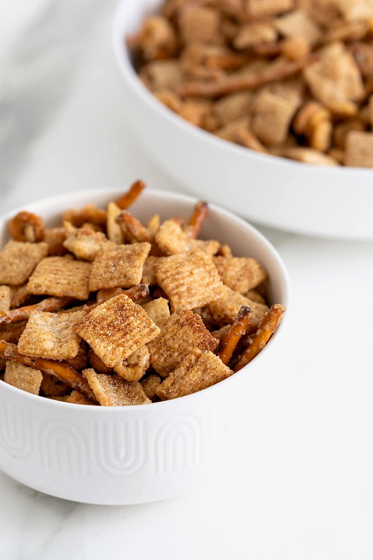 Two white bowls of cinnamon cereal snack mix with pretzels.