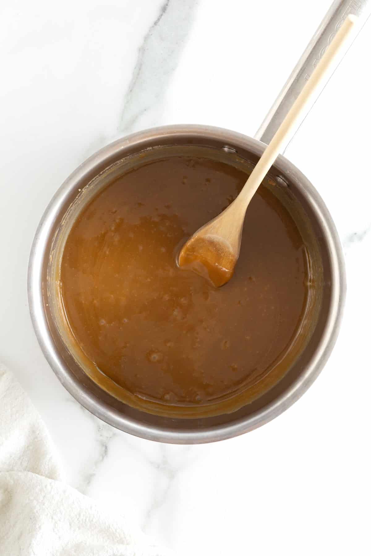 A wooden spoon in a light metal saucepan of caramel sauce.