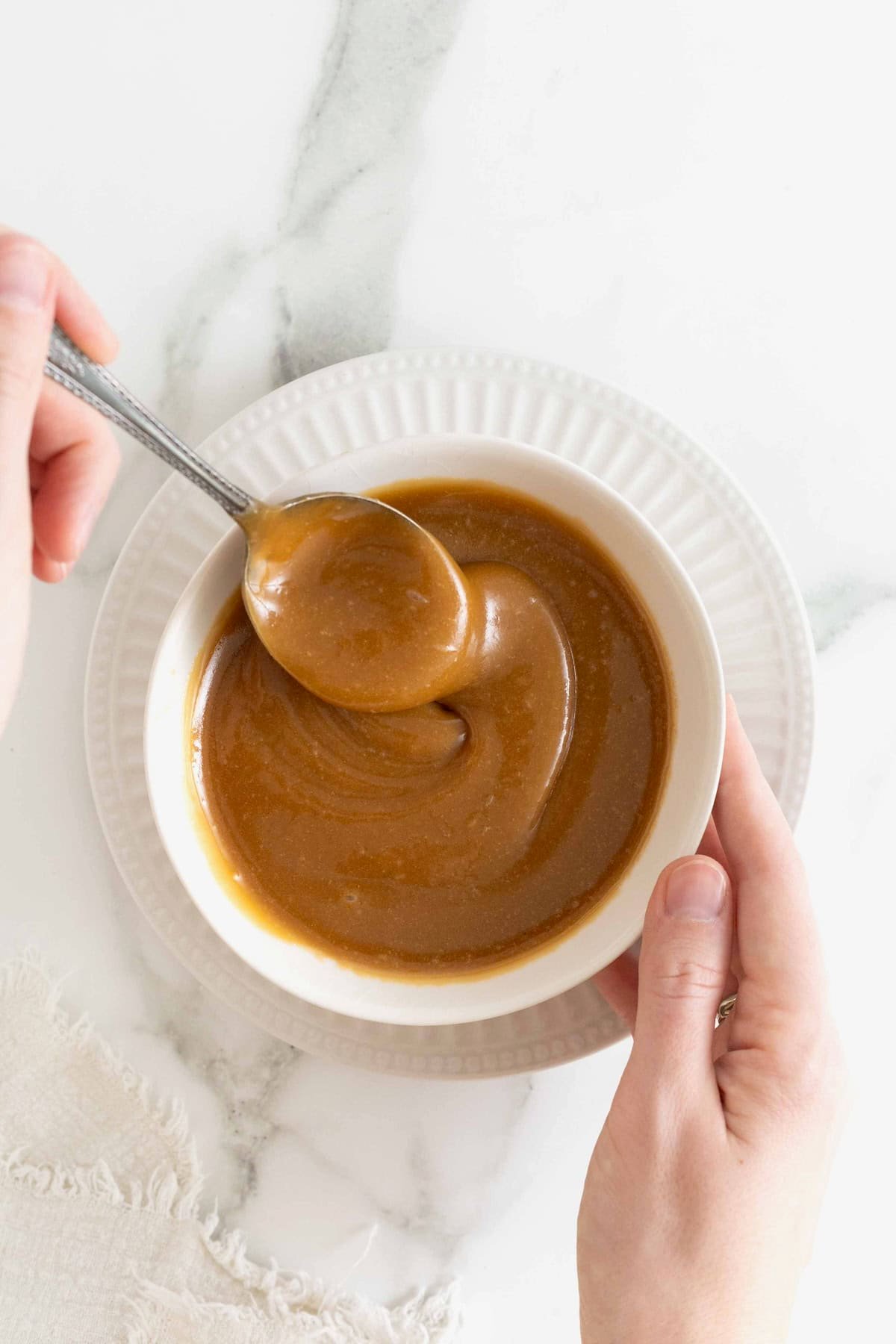 Caramel sauce in a white bowl being stirred with a metal spoon.
