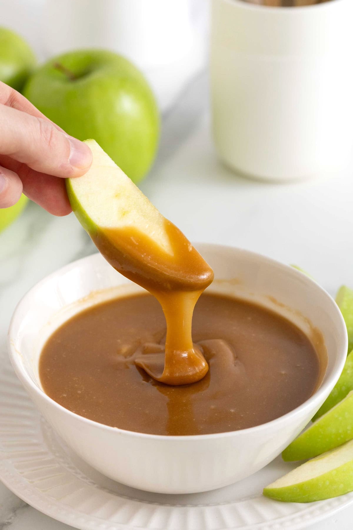 A green apple slice being dipped into a small white bowl of caramel.