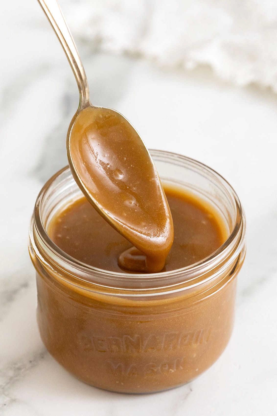 A spoon being lifted from a short wide-mouth mason jar of caramel sauce.