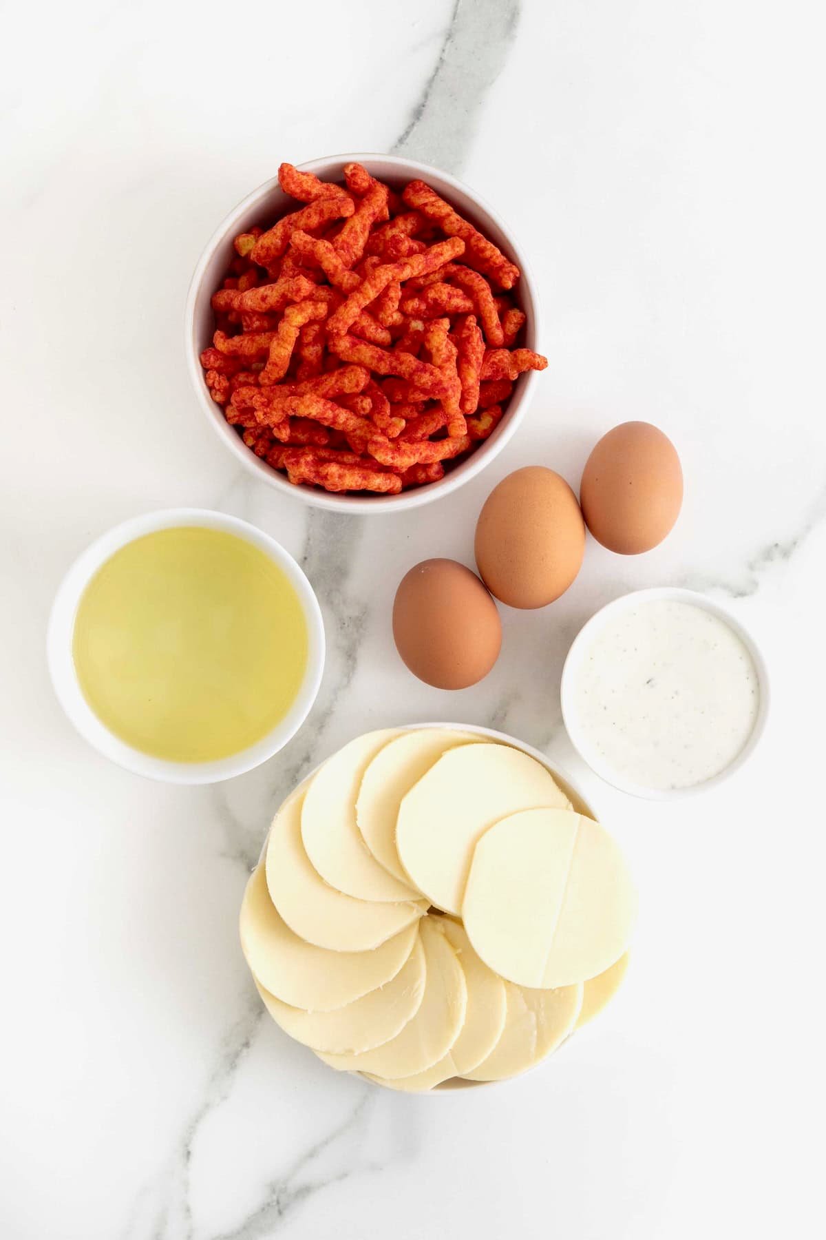 Ingredients to make Flamin' Hot Cheeto Mozzarella Hearts in small white dishes on a white marble counter.