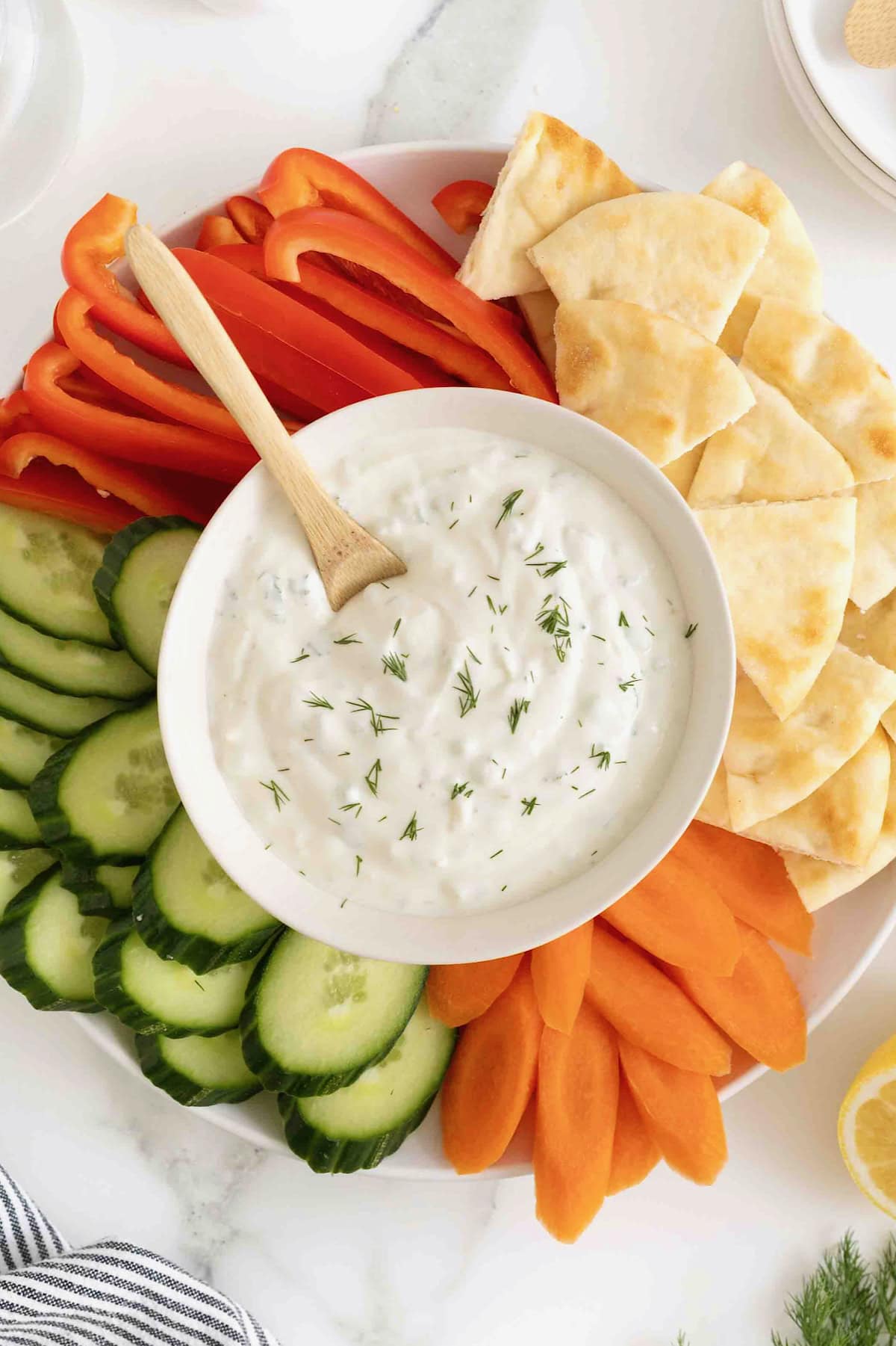 A white bowl of tzatziki sauce surrounded by slices of cucumber red pepper, carrot and pita wedges.