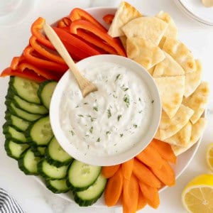 A white bowl of tzatziki sauce surrounded by slices of cucumber red pepper, carrot and pita chips.