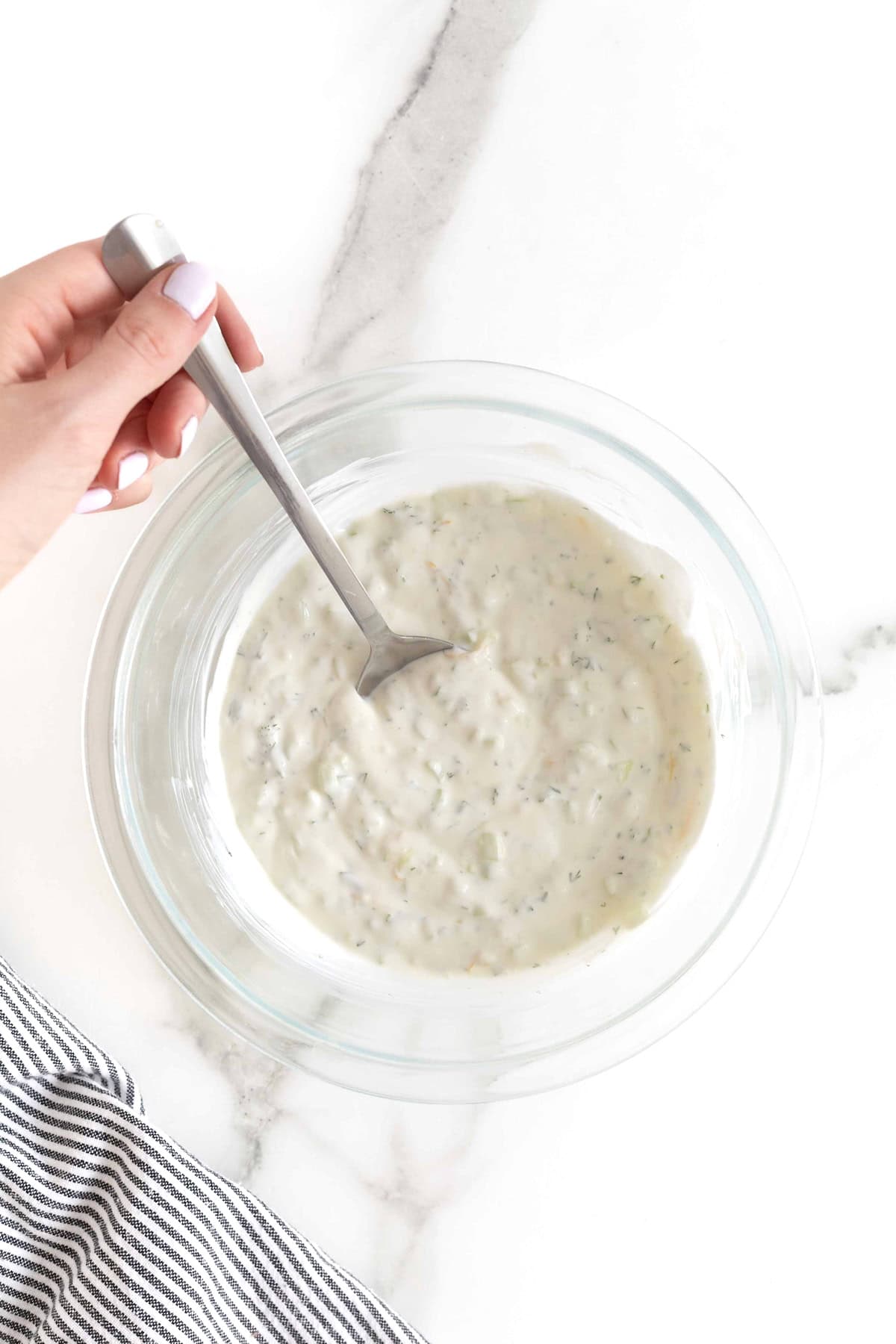 A hand stirring a glass mixing bowl of tzatziki sauce.