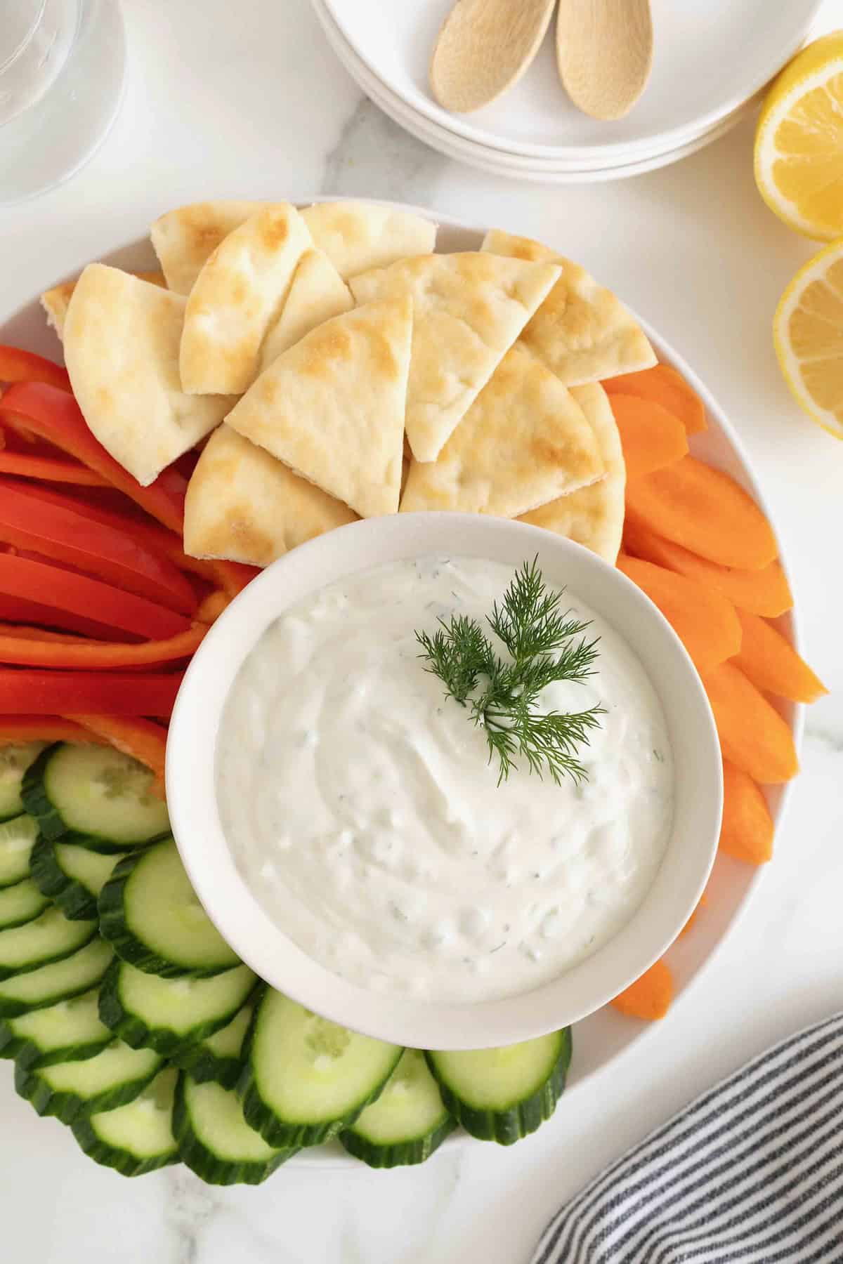 A white bowl of tzatziki sauce surrounded by slices of cucumber red pepper, carrot and pita chips. There is a sprig of fresh dill on the dip.