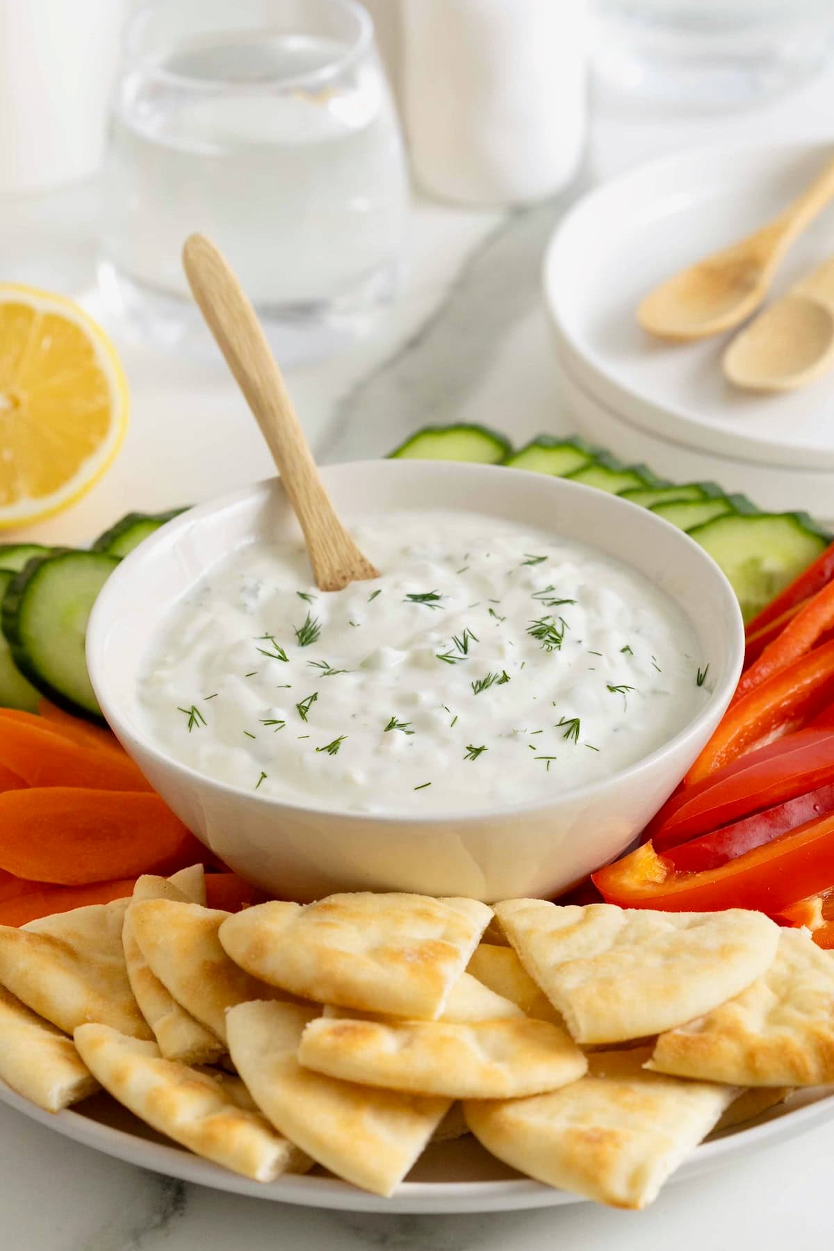 A wooden spoon in a white bowl of tzatziki sauce surrounded by pita wedges and vegetables.