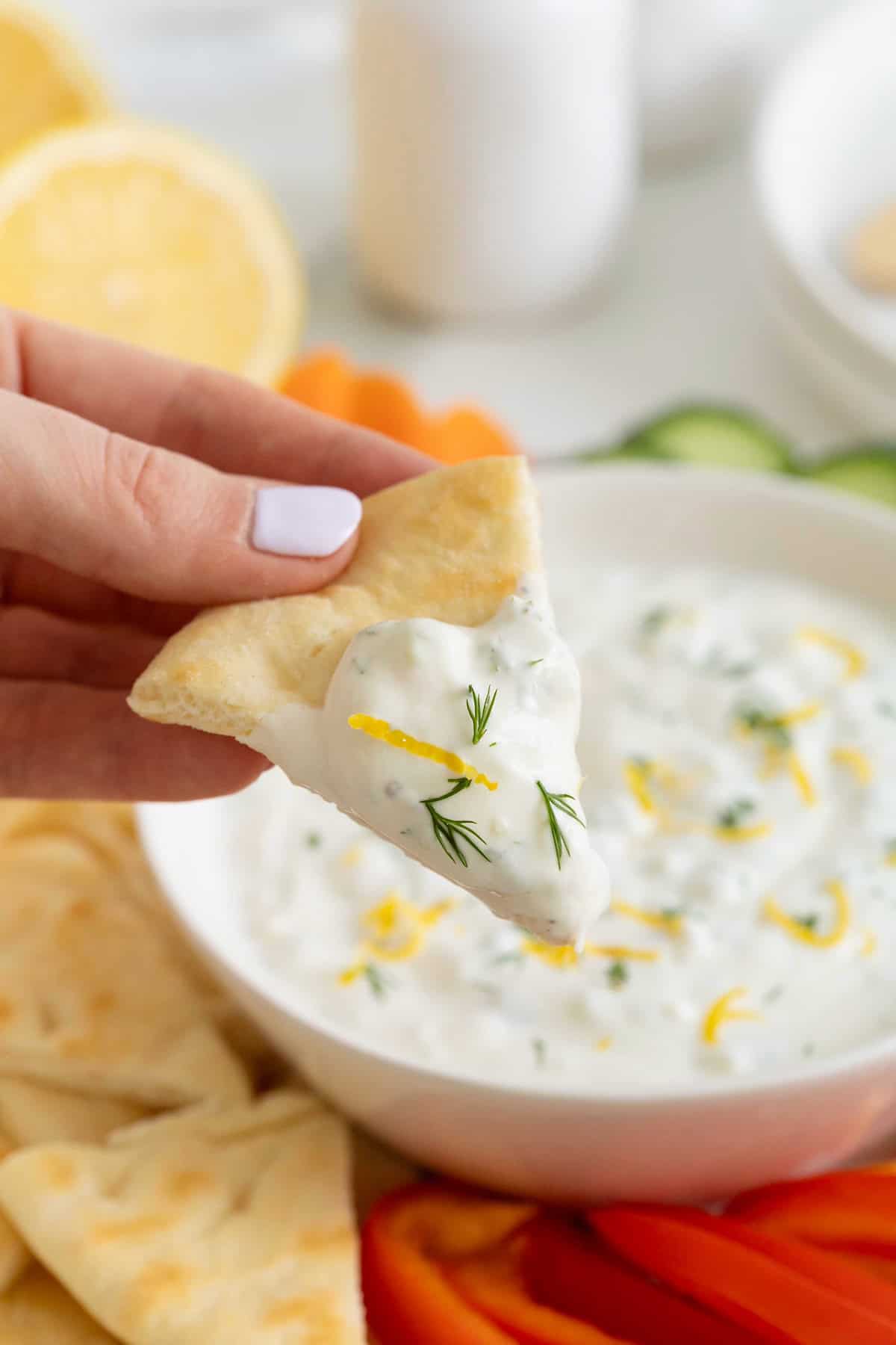 A hand holding a wedge of fresh pita dipped in tzatziki sauce.
