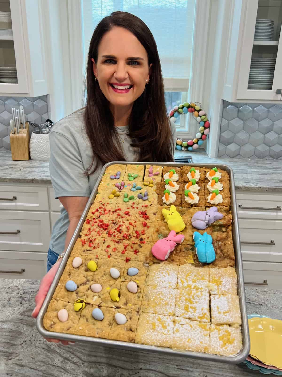 Maegan Brown wearing a short sleeved gray top holding a sheet pan of Easter cookie bars.
