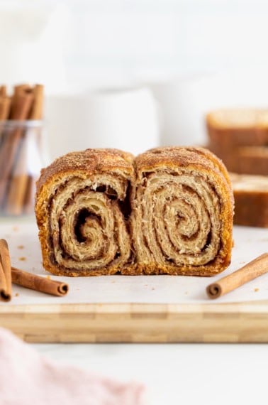 A cinnamon sugar crescent roll loaf on a light wood cutting board. There are cinnamon sticks scattered around it.