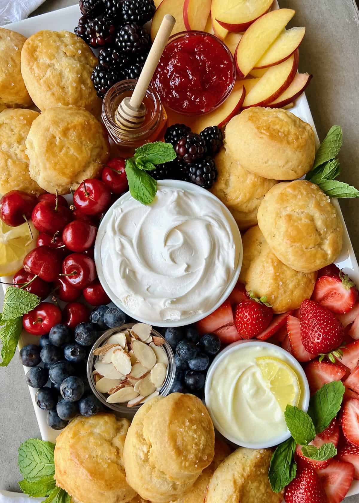 A food board piled with scones, fresh fruits, and small dishes of creamy toppings.