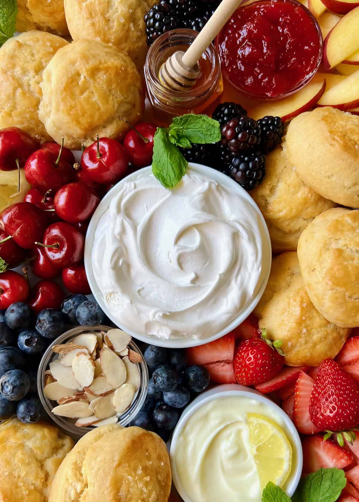 A bowl of whipped cream surrounded by fresh cherries, blueberries, strawberries and scones.