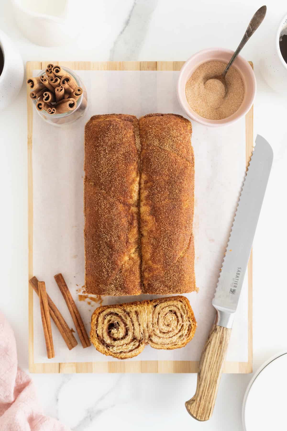 A cinnamon sugar crescent roll loaf on a light wood cutting board. There are cinnamon sticks scattered around it.