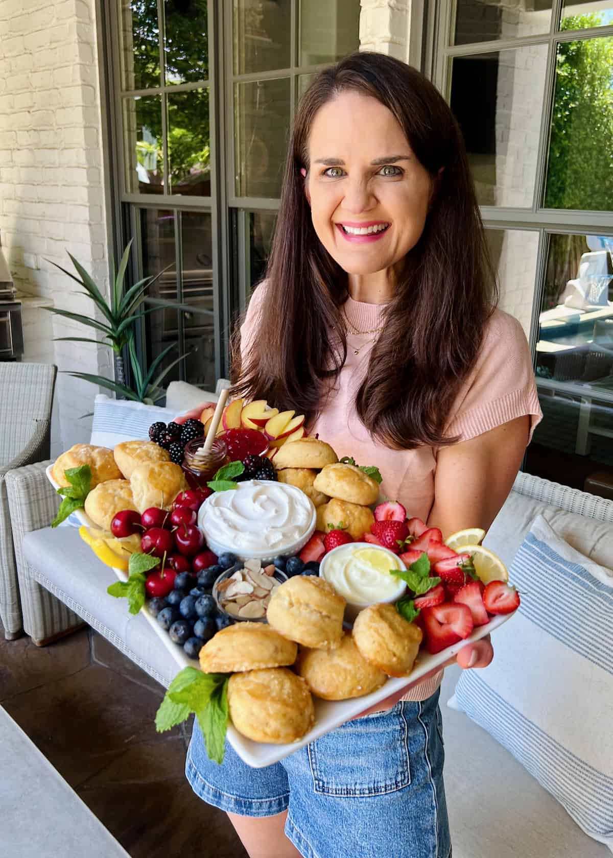 Maegan Brown wearing a light pink t-shirt holding a platter of scones, fresh fruit, whipped cream and honey.