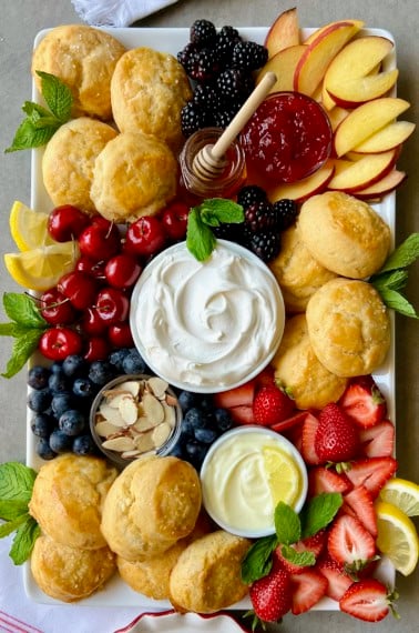 A food board piled with scones, fresh fruits, and small dishes of creamy toppings.
