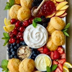 A rectangular white serving platter piled with scones, fresh fruits, and small dishes of creamy toppings.