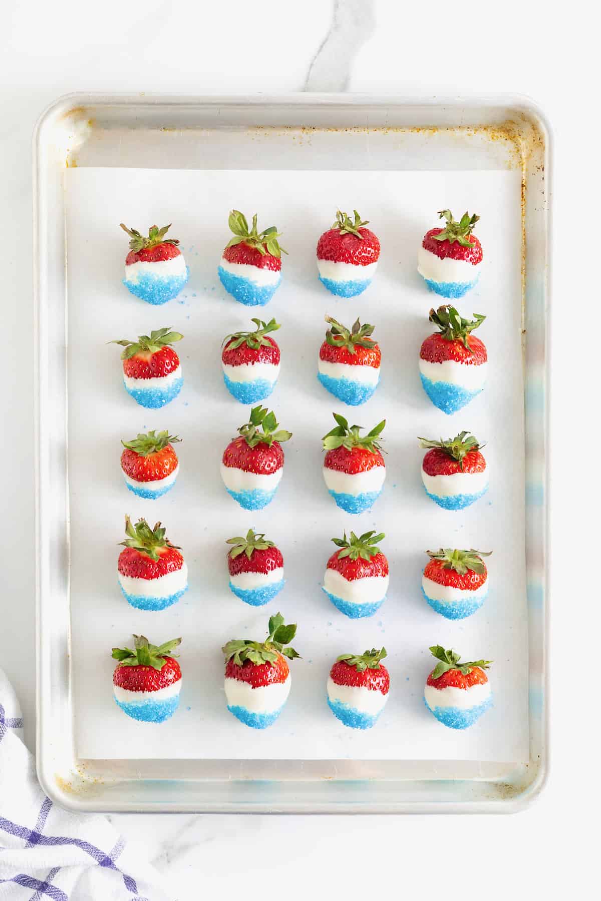 Twenty strawberries dipped in white candy coating and blue decorating sugar on a parchment lined baking sheet.