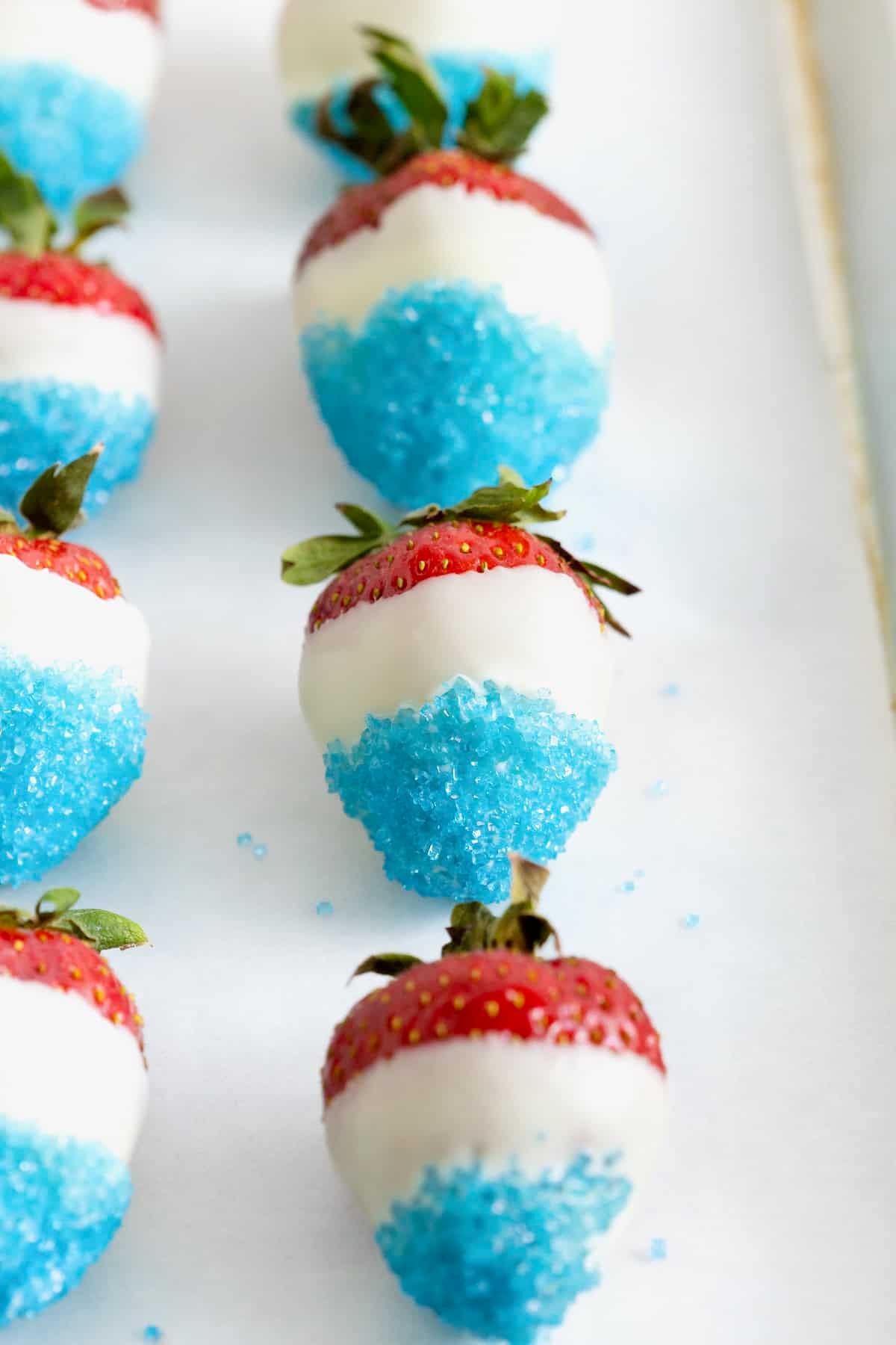 Seven strawberries dipped in white candy coating and blue decorating sugar on a parchment lined baking sheet.