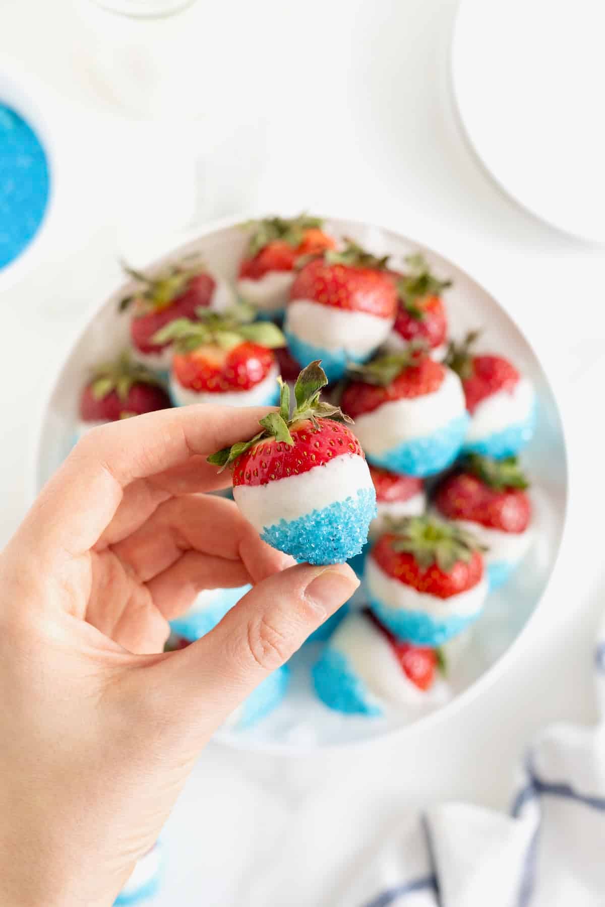 A hand holding a red white and blue strawberry over a platter of strawberries dipped in candy coating and decorating sugar.
