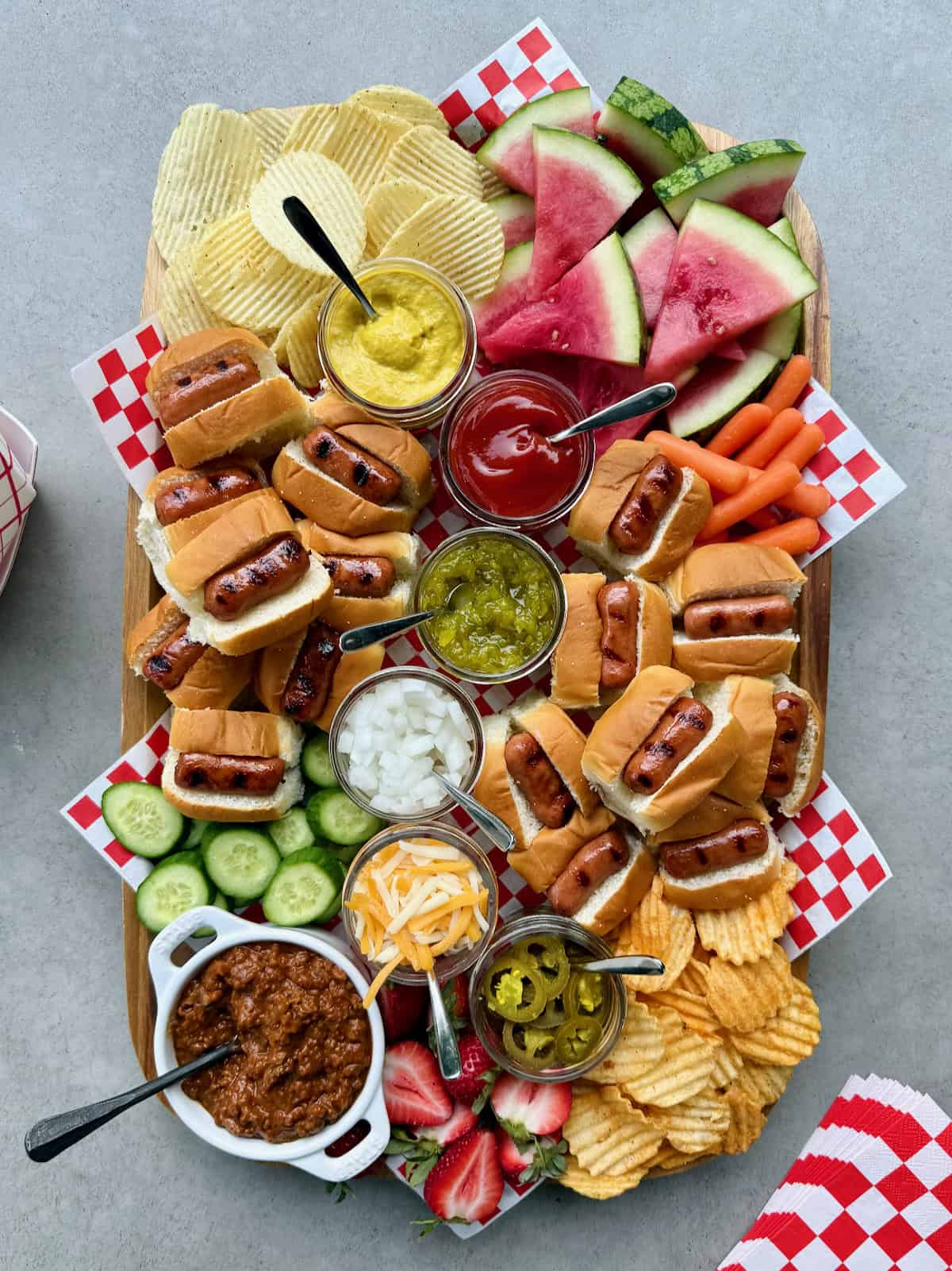 A rectangular foo board loaded with mini hot dogs and fixings, chips, and watermelon wedges.