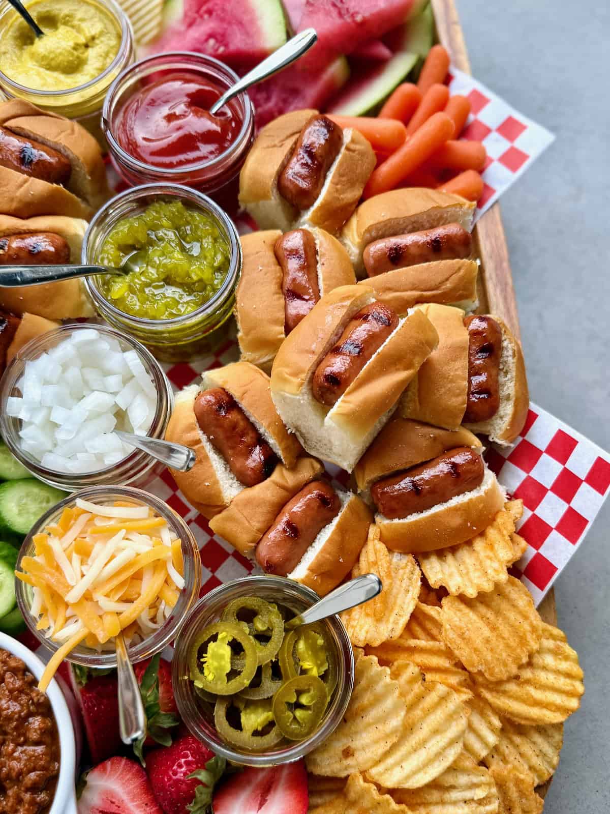 A rectangular foo board loaded with mini hot dogs and fixings, chips, and watermelon wedges.