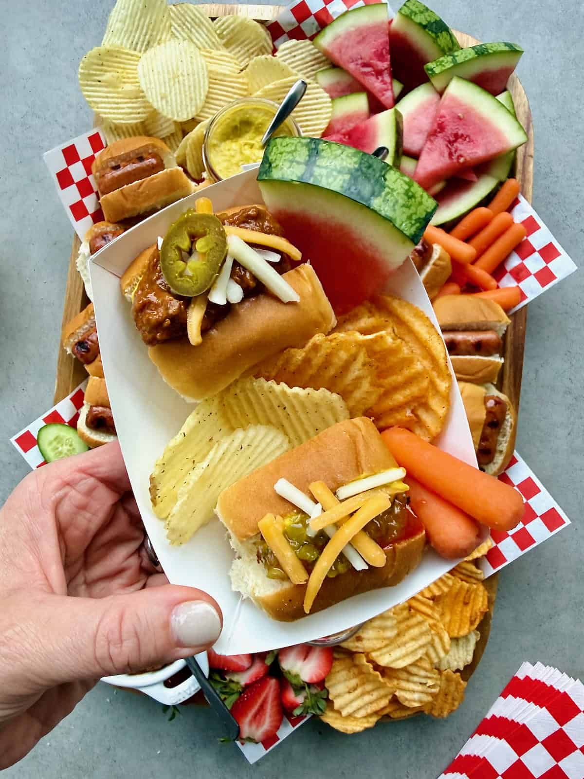 Two mini hot dogs surrounded by chips, a watermelon wedge and baby carrots. 