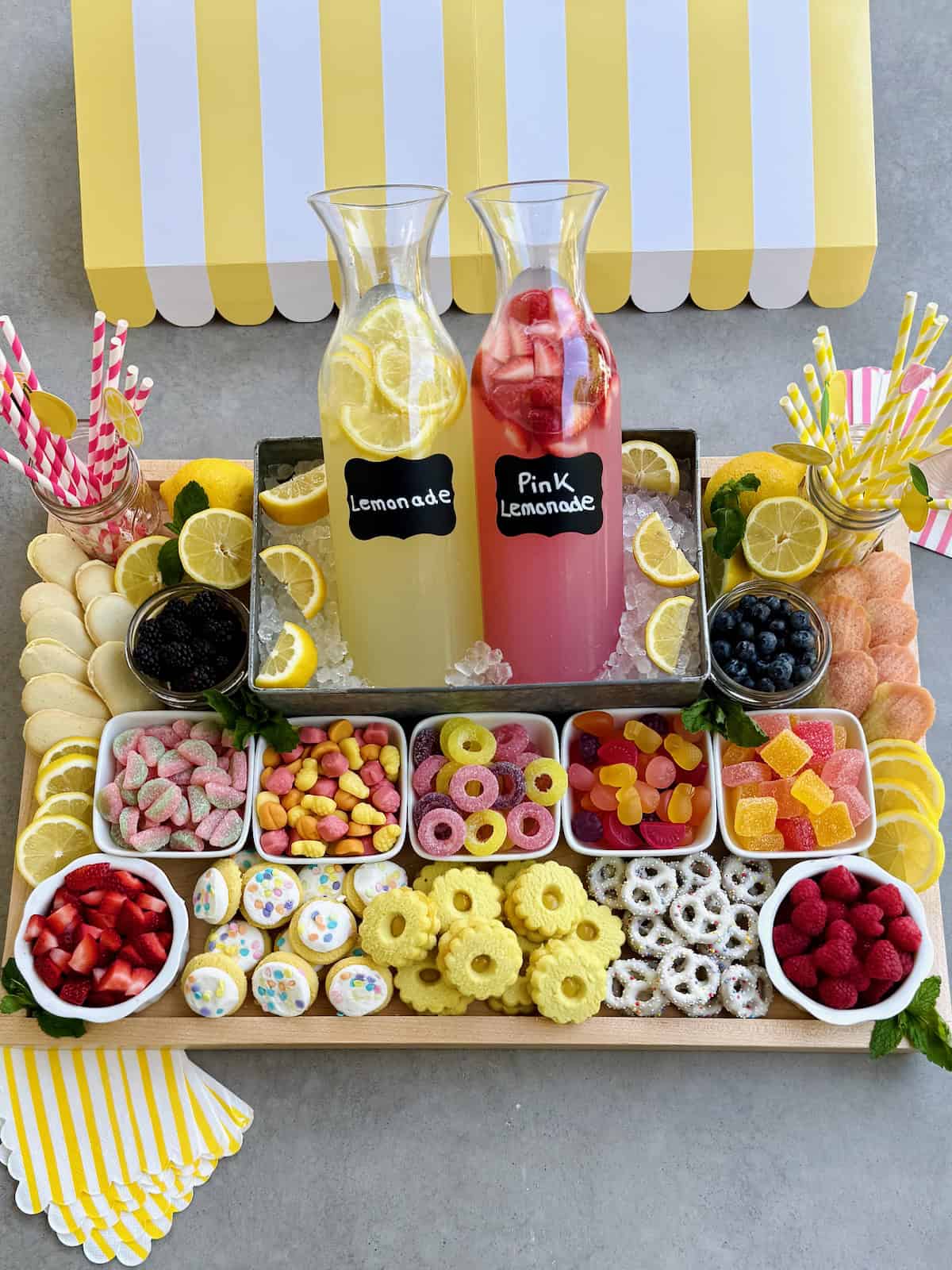 A food board with lemonade options and snacks, including gummy candies, cookies and chocolate dipped pretzels.