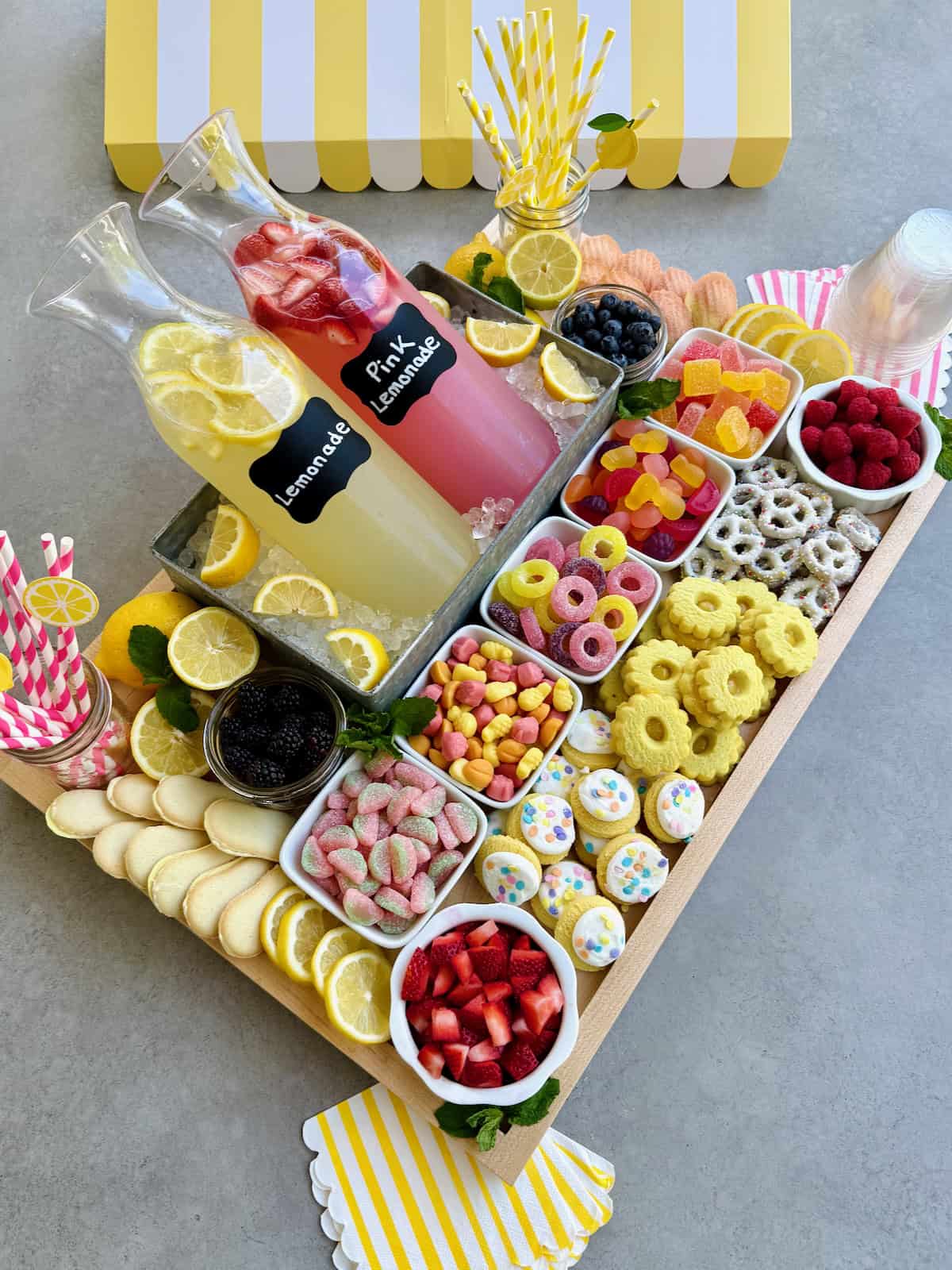 A food board with lemonade options and snacks, including gummy candies, cookies and chocolate dipped pretzels.