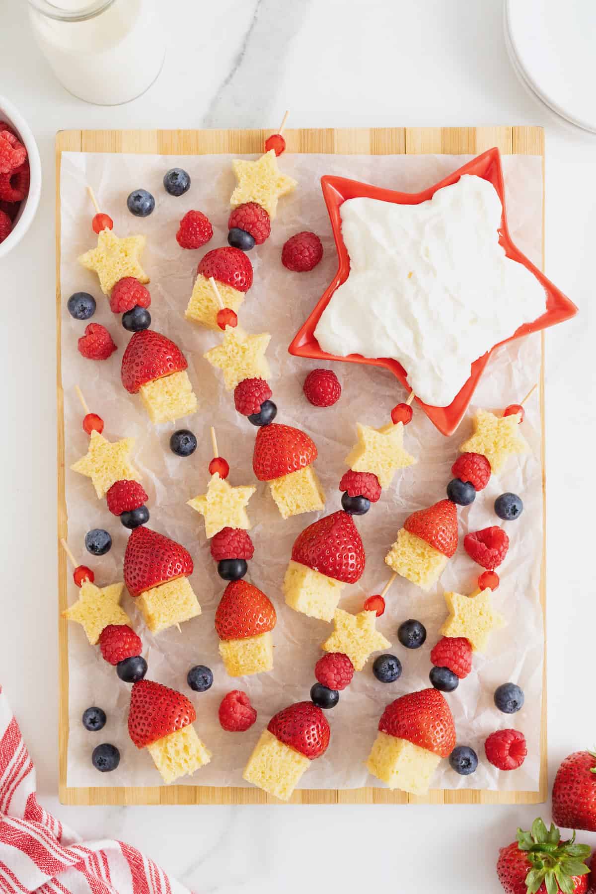 A parchment lined wood board filled with berry and shortcake skewers. There is a red, star-shaped dish of whipped cream at the top right of the board.