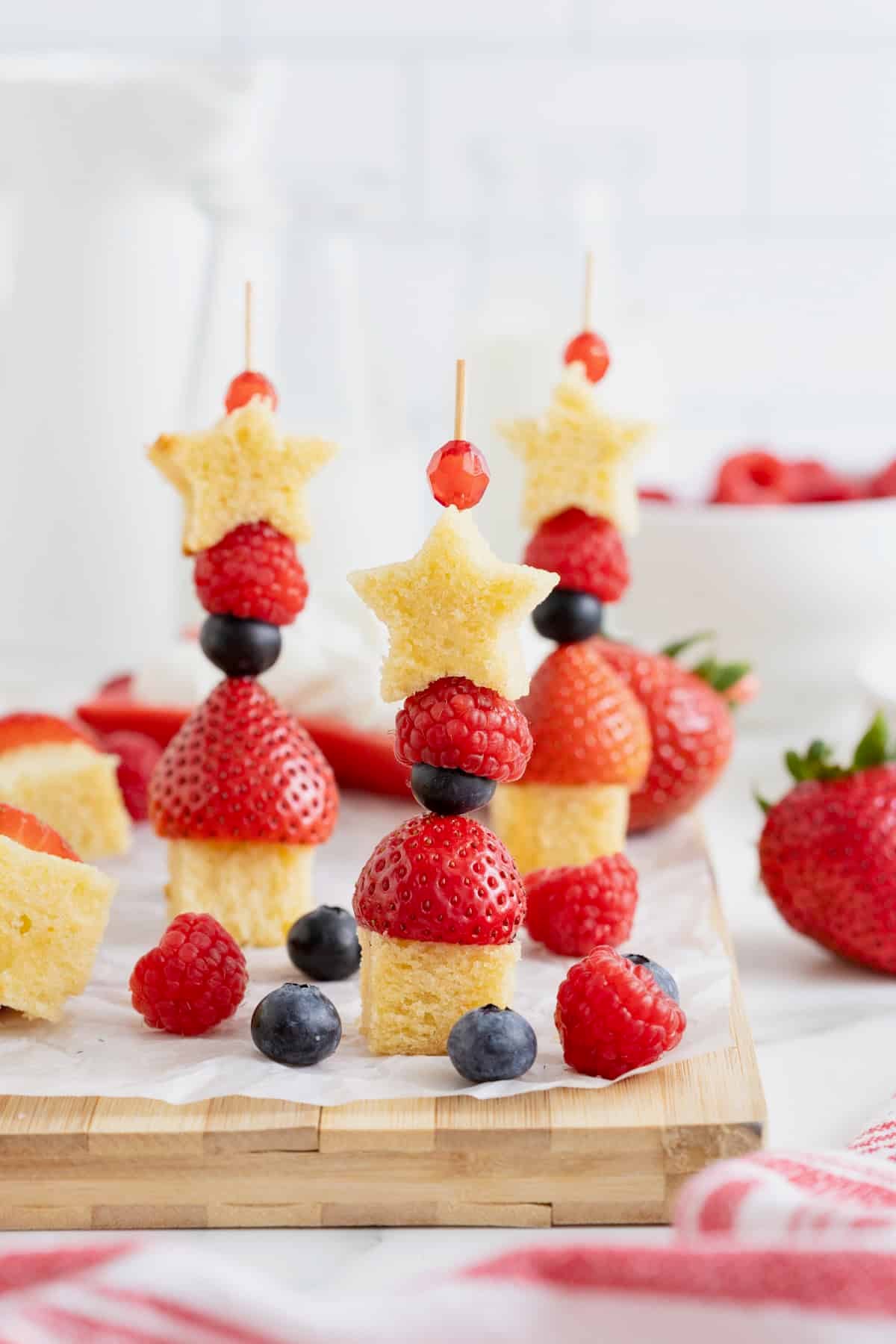 Three skewers made of poundcake cubes, strawberries, blueberries, raspberries and a poundcake star. In the background is a white bowl of blueberries and a red dish of whipped cream. The skewers are surrounded by berries.