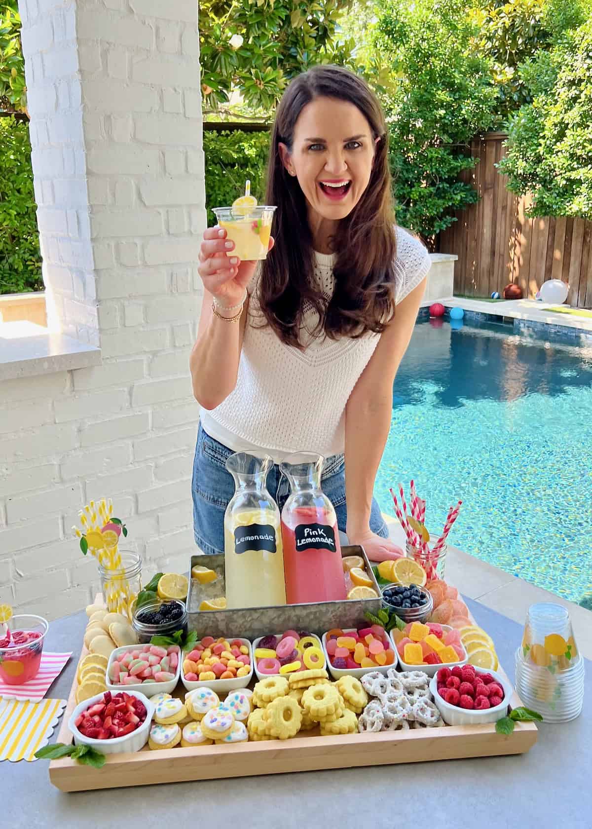 Maegan Brown wearing a white blouse and holding a cup of lemonade. In front of her is a food board with candies, cookies and lemonade.