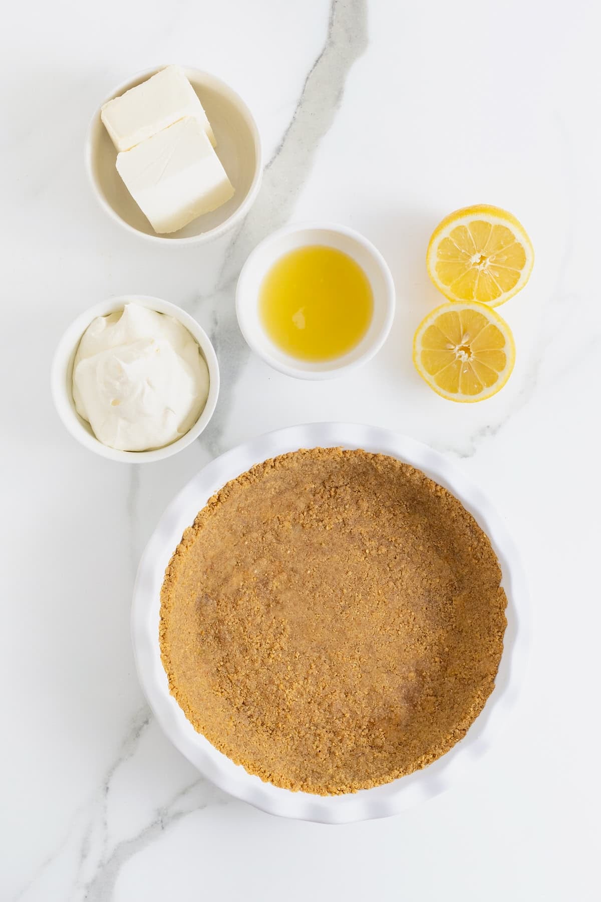 Ingredients to make frozen lemonade pie in white dishes on a white marble counter.