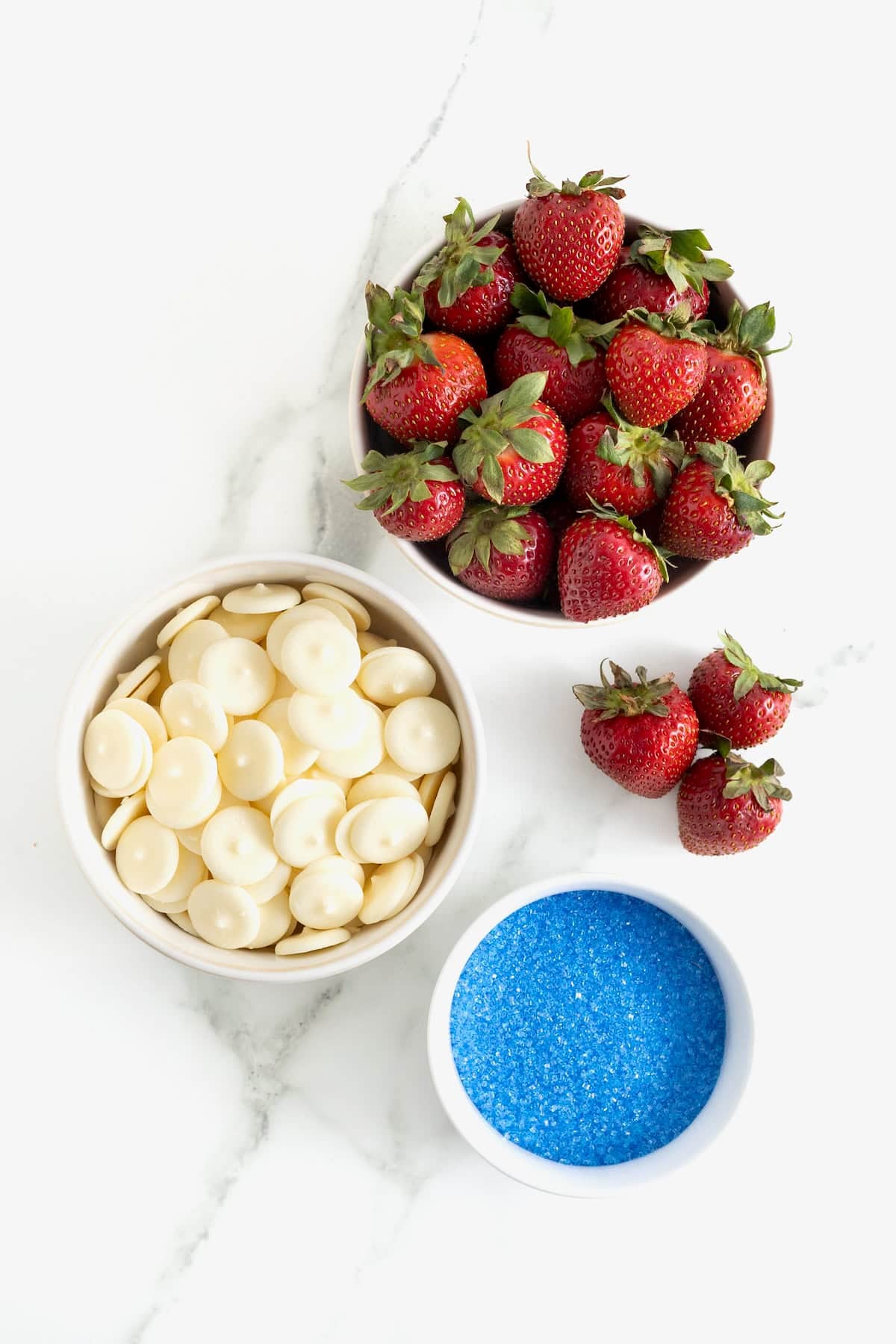 Ingredients to make red, white and blue strawberries in white dishes on a white marble counter.