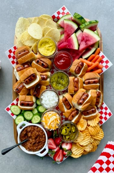 A rectangular foo board loaded with mini hot dogs and fixings, chips, and watermelon wedges.