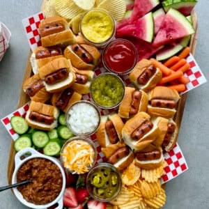 A rectangular foo board loaded with mini hot dogs and fixings, chips, and watermelon wedges.
