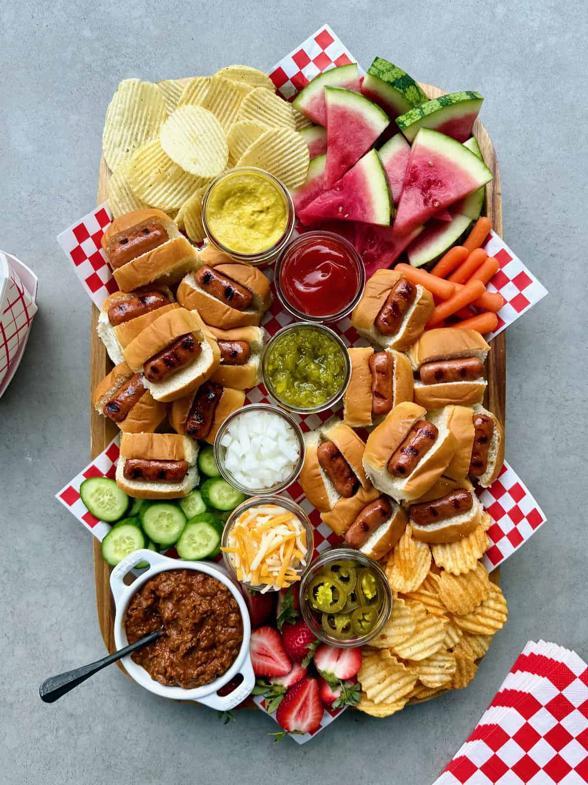 A rectangular foo board loaded with mini hot dogs and fixings, chips, and watermelon wedges.