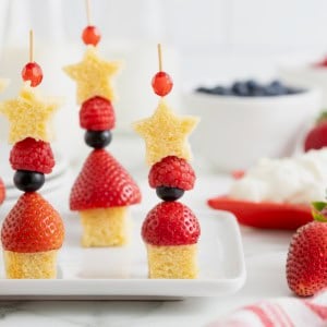 Three skewers made of of shortcake cubes, strawberries, blueberries, raspberries and a shortcake star. In the background is a white bowl of blueberries and a red dish of whipped cream.