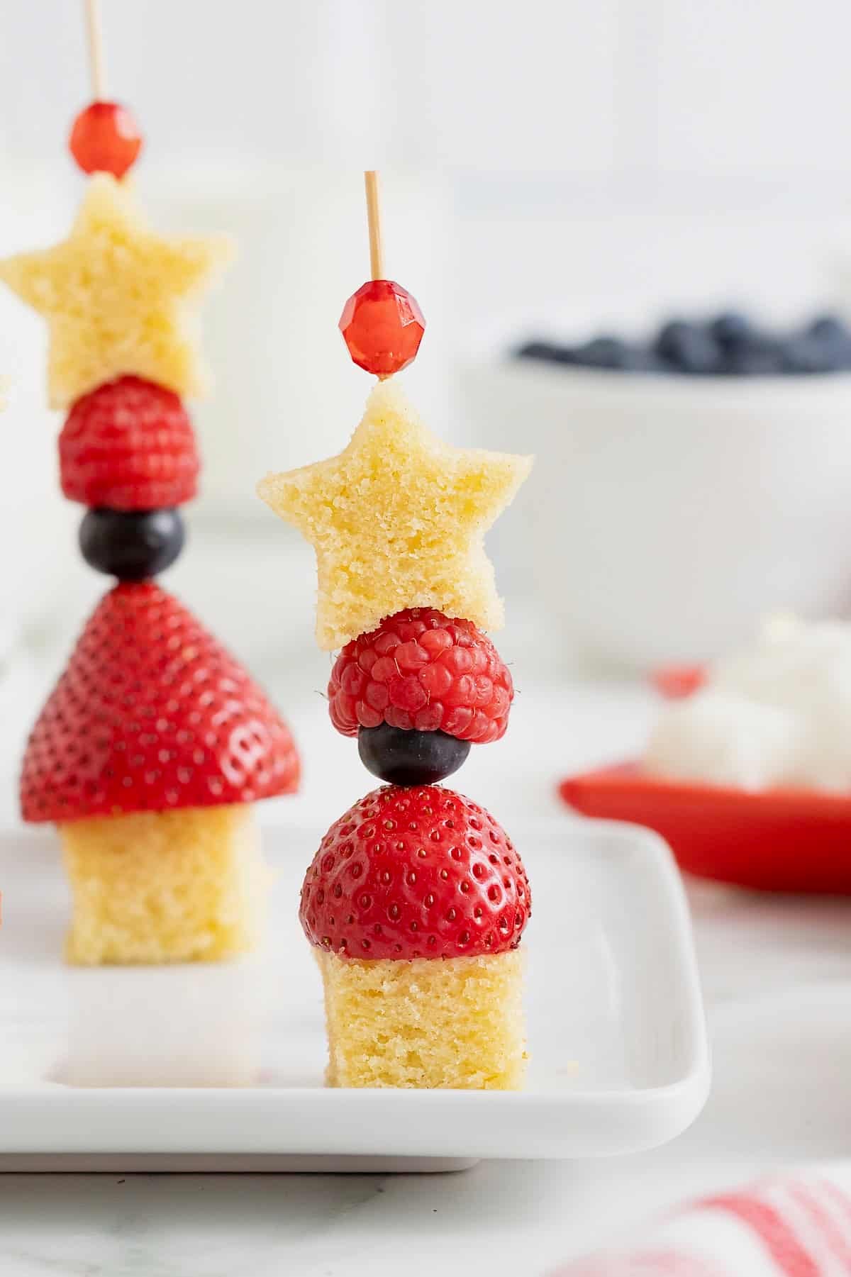 Two skewers made of of shortcake cubes, strawberries, blueberries, raspberries and a shortcake star. In the background is a white bowl of blueberries and a red dish of whipped cream.