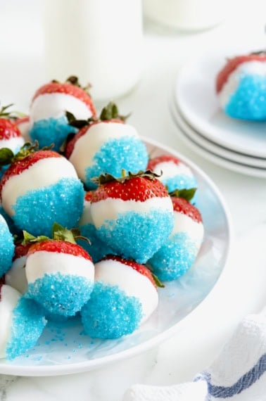 A white platter piled with strawberries decorated with white candy coating and blue decorating sugar.