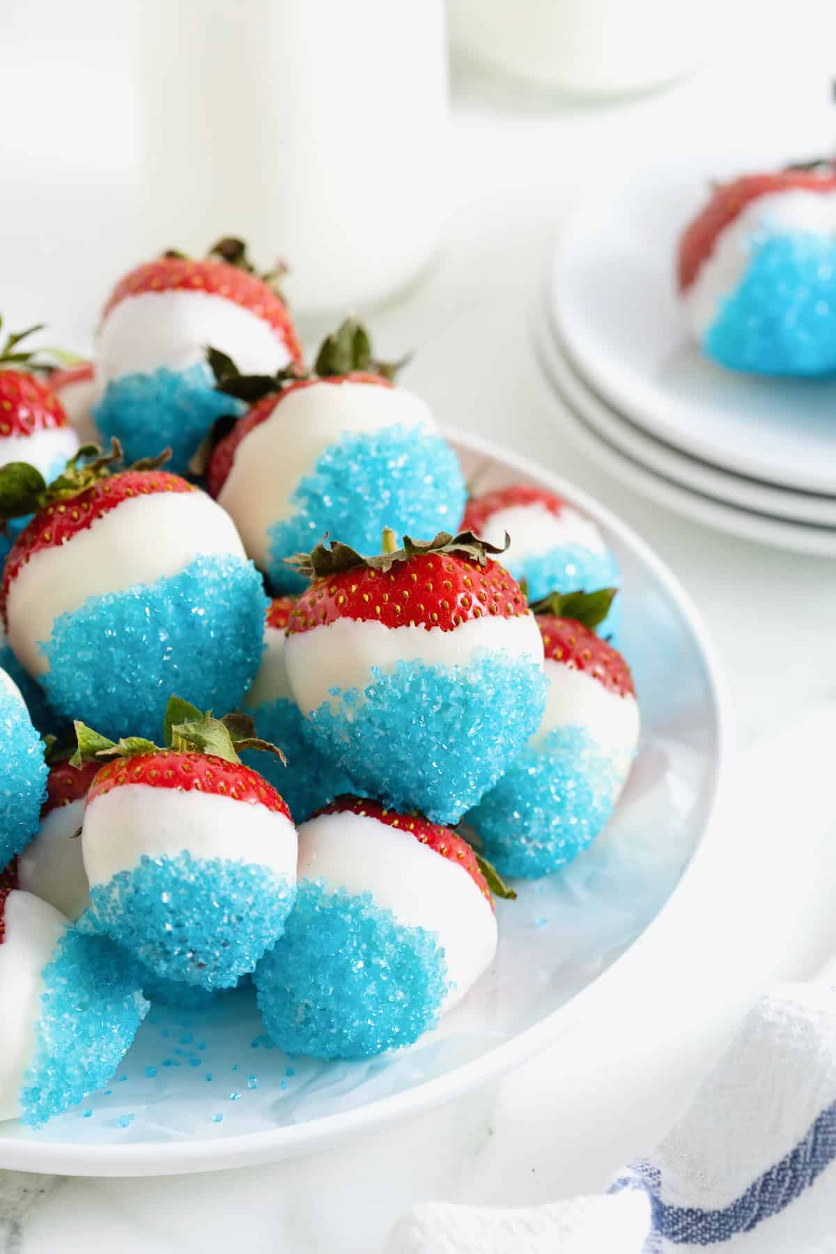 A white platter piled with strawberries decorated with white candy coating and blue decorating sugar.