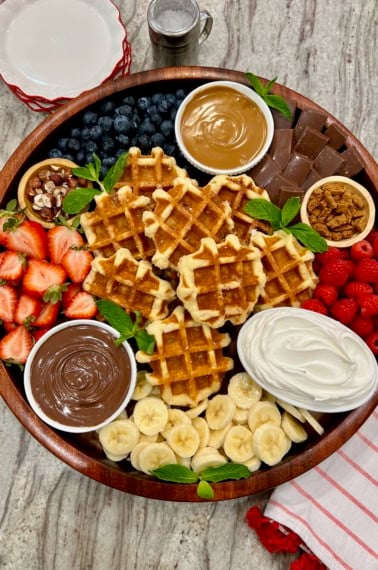 A round wood food tray loaded with Belgian waffles, fruit, Nutella, biscoff butter and whipped cream.
