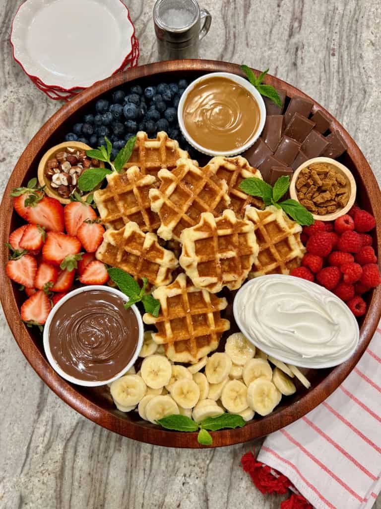 A round wood food tray loaded with Belgian waffles, fruit, Nutella, peanut butter and whipped cream.