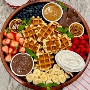 A round wood food tray loaded with Belgian waffles, fruit, Nutella, biscoff butter and whipped cream.
