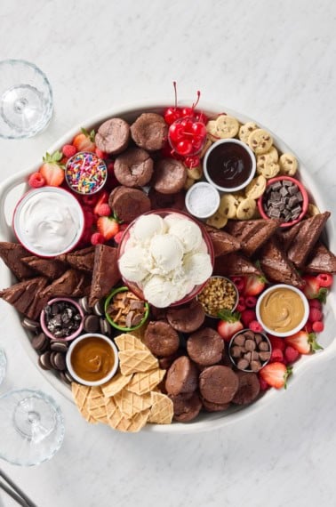 A large round serving tray filled with brownie bites, brownie triangles, scoops of vanilla ice cream, and various toppings.