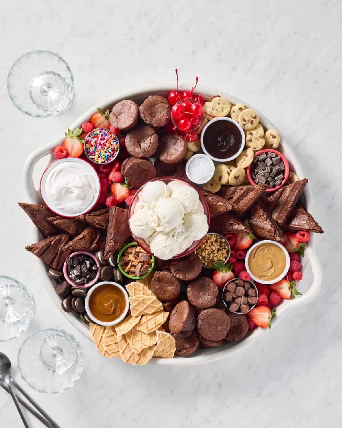 A large round serving tray filled with brownie bites, brownie triangles, scoops of vanilla ice cream, and various toppings.