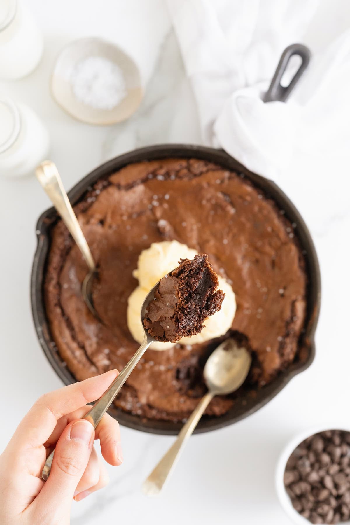 A skillet brownie topped with ice cream. There are two spoons in the skillet and one lifting a bite of brown away.
